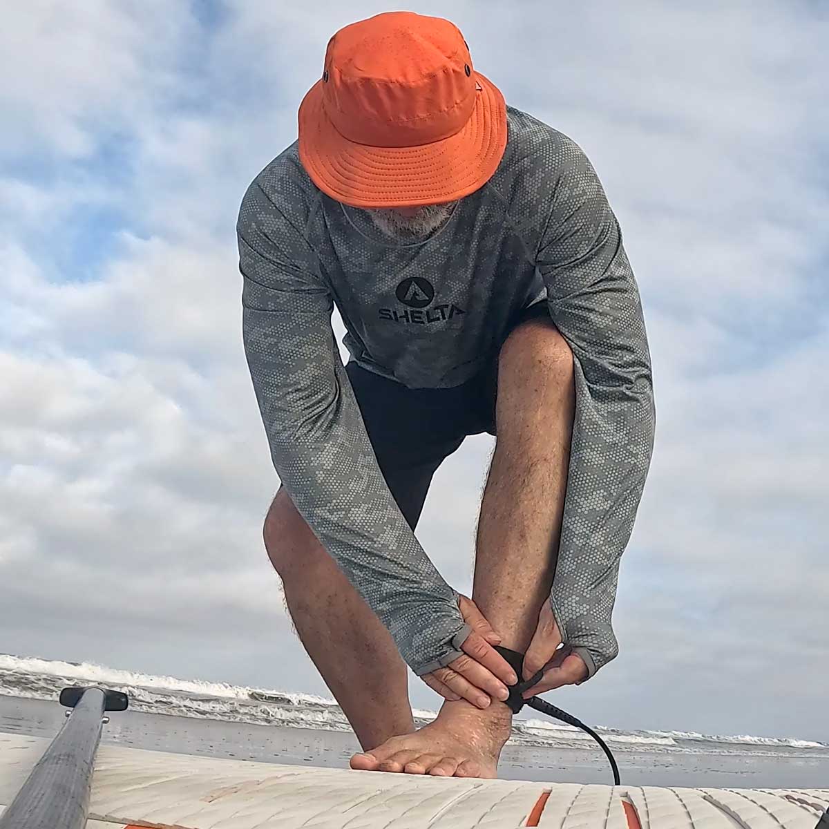 picture of a man putting on his surfboard leash wearinga shelta sun hat and aquaterra sun shirt